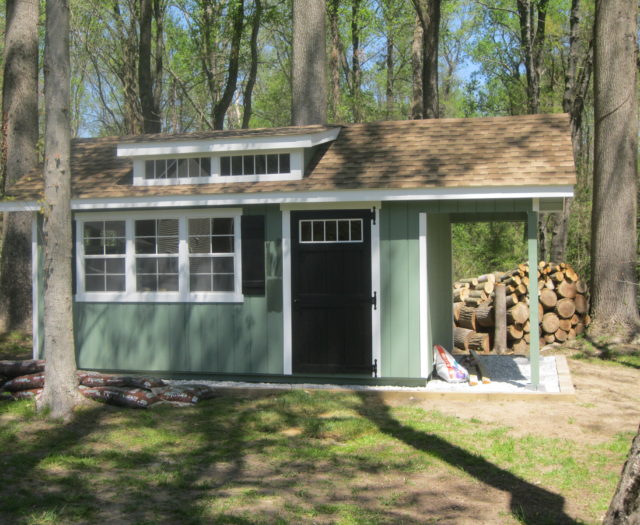 green classic studio dormer shed with pile of wood in back